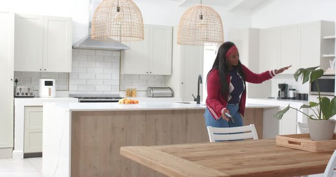 African American woman walking in kitchen raising phone and lifting cup cueing to plant, copy space