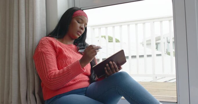 African American woman in coral sweater writing on brown pad on window seat, light inspiring noting