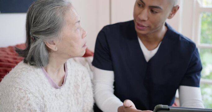 African aide in scrubs placing hand, speaking, assisting on tablet on sofa, senior in cream sweater