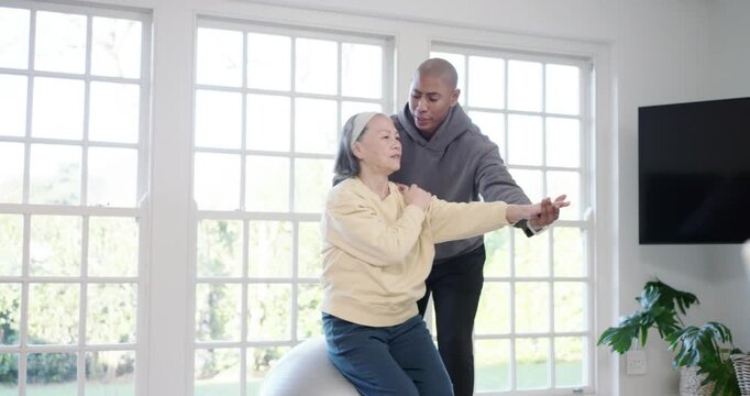 Senior Asian woman on exercise ball caregiver placing hand guiding arm for mobility in living room