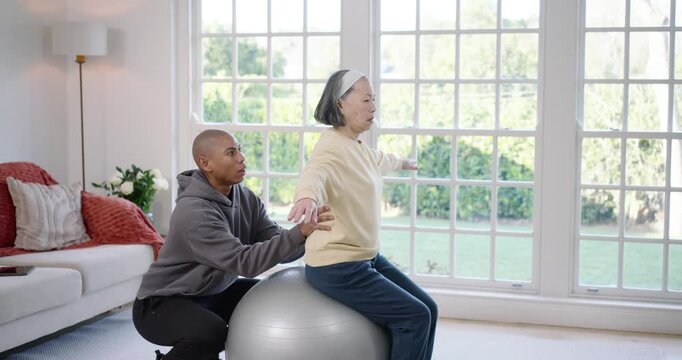Senior Asian woman, African American PT steadying silver ball, raising arms for balance at home