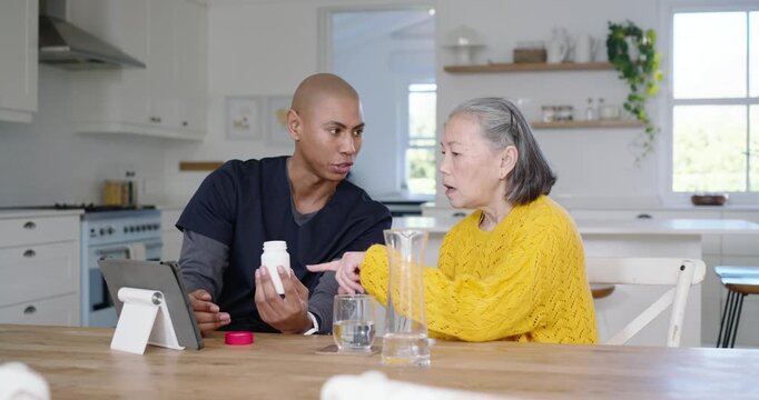 Caregiver in scrubs showing pill-label on tablet to Senior Chinese woman at kitchen explaining meds