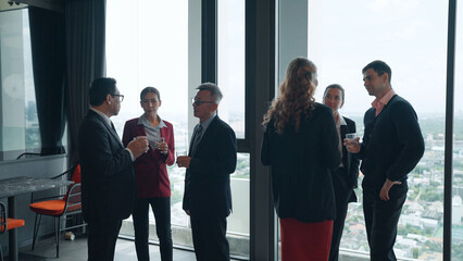Business executives hold drinks and converse in small groups near floor-to-ceiling windows...