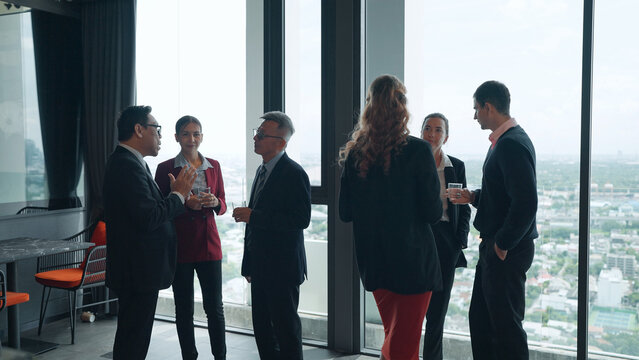 A diverse group of business professionals in formal attire network and chat in small groups by floor-to-ceiling windows overlooking a city skyline