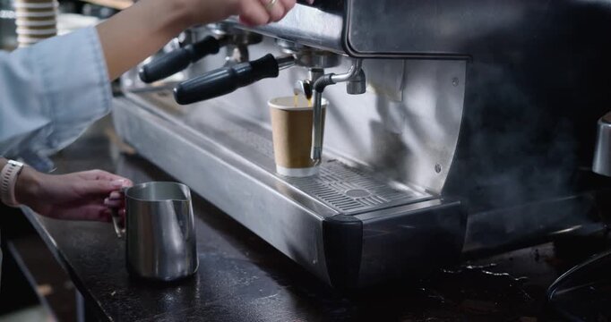 Barista is adjusting machine lever, espresso is extracting into takeaway cup and milk is steaming