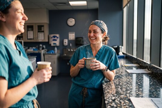 Two smiling female doctors in scrubs taking a coffee break and talking together in hospital breakroom