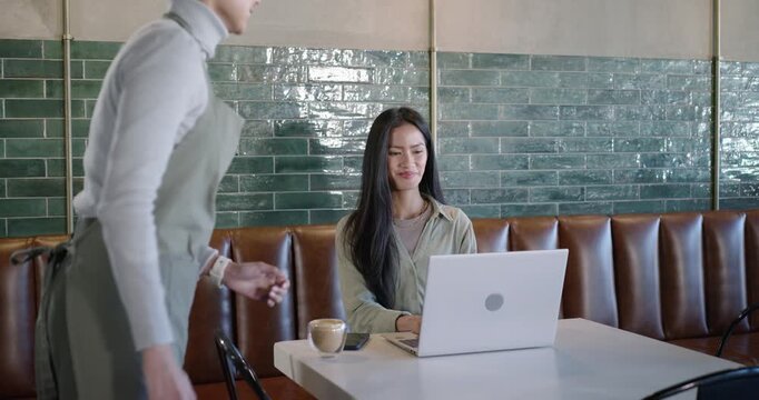 Mid-adult Asian woman working on laptop at cafe, server placing coffee cup, pausing, smiling