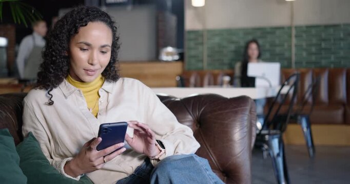 Camera dollying in, mid-adult Asian woman scrolling and replying on blue smartphone at cafe sofa