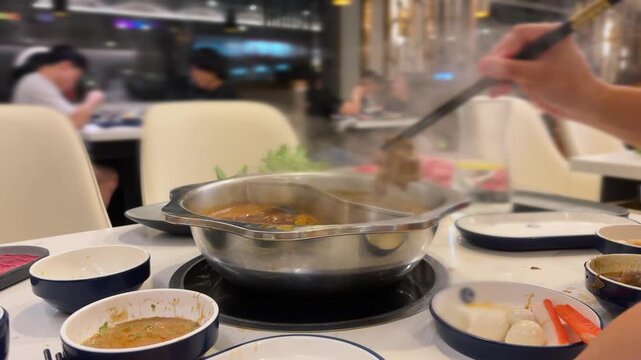 A traditional two-flavor hot pot with simmering broths boils at a restaurant table in Toronto, Canada, with a person using chopsticks to dip sliced beef and mutton into the steaming soup.