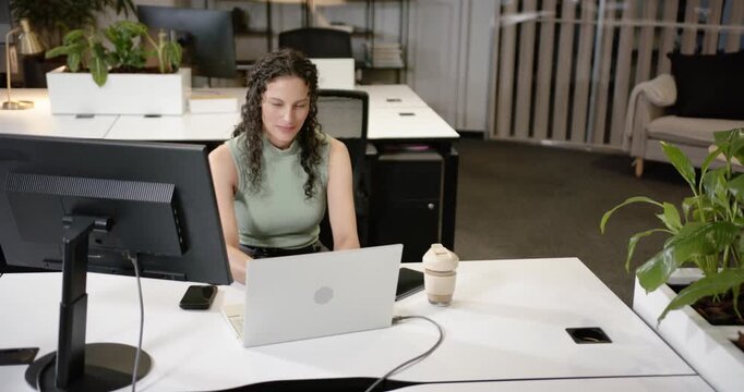 African woman opening laptop, typing and glancing at monitor while working at shared office desk