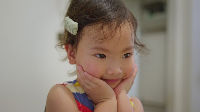 A cute Asian toddler girl holds her face with both hands and smiles gently, looking away with a sweet expression of childhood wonder and contentment in her home.