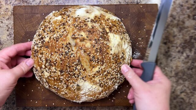 Top-down view of female hands slicing a round loaf of homemade sourdough bread covered with sesame and flax seeds in half on a wooden board.