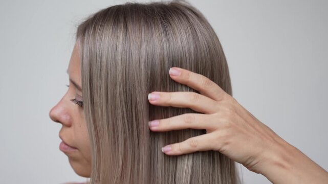 Close-up of young woman running fingers through smooth ash blond hair, demonstrating silky texture, salon coloring, airtouch highlights and keratin straightening effect on a light background
