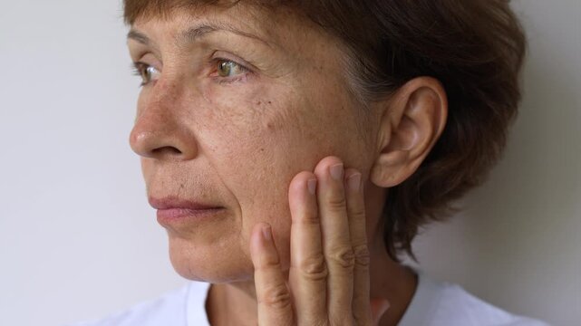 Mature woman touching skin around her eye and cheek, showing crow&rsquo;s feet, wrinkles and visible signs of aging on a light background
