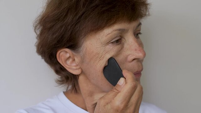 Close-up of mature woman massaging her cheek with a gua sha scraper for lifting, relaxation and facial skincare on a light background