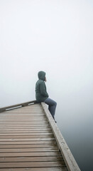 Fototapeta premium A person in a hooded jacket sits on a wooden pier, looking into the dense fog symbolizing contemplation and isolation