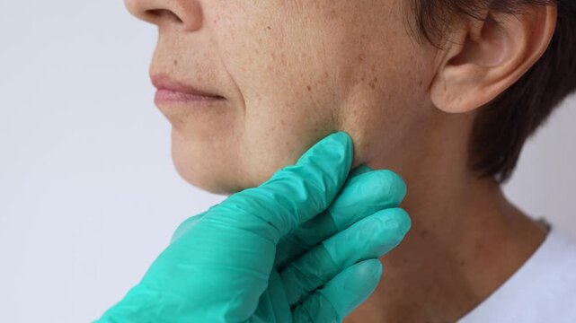 Gloved doctor&rsquo;s hand palpating the lower face and jawline of a mature woman during a medical examination and preoperative assessment on a light background 