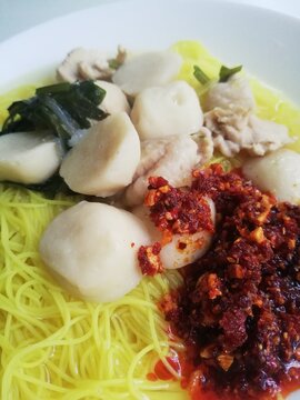 Close-up shot of rice vermicelli noodle soup topped with fishballs and spicy chili oil, served on a white plate. 