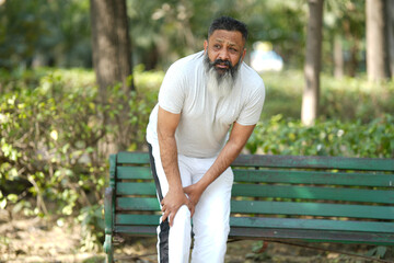 Fototapeta premium Indian senior man suffering from knee pain at park. Pain In The Elderly, Health care. Bearded man painful on knee.