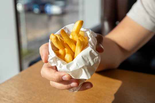 Man holding paper package of golden fried potato. Male hand with french fries in fast food restaurant. Street food snack and takeaway meal. Tasty fried potato concept for cafe menu and food delivery.
