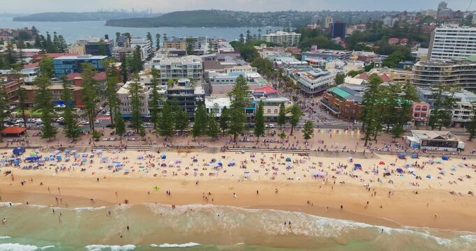 Manly Beach in Sydney, Australia, showing crowds of people enjoying the sand and ocean, with the vibrant beachfront cityscape, hotels, and businesses lining the popular coastal promenade on summer day