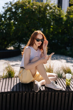 Vertical portrait of stylish ginger woman in casual summer clothes sitting on park bench using smartphone. Bright sunlight creating relaxed mood. Modern lifestyle and technology concept.