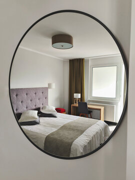 Oval wall mirror reflecting modern bedroom with double bed, upholstered headboard, window, desk and soft neutral decor