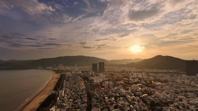 Aerial timelapse of Quy Nhon, Vietnam, transitioning from twilight through sunset into night. The city and sea stretch across the landscape, with mountains and drifting clouds in the background as