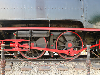 Red wheels turn on dark steel train car. Rust and wear mark historic locomotive frame. Chain links connect moving parts with strength. Trackbed stones lie beneath heavy machinery © Erwin Eka Chendra