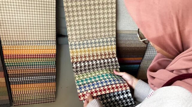 Young Asian Muslim woman in hijab examining and choosing fabric swatches and color samples for interior design project in a shop.