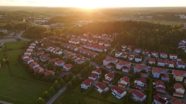 Aerial drone footage of a peaceful neighborhood with compact homes and red roofs surrounded by green vegetation and forest during golden sunset