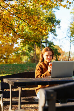 Mid-adult woman watching educational video on laptop with takeaway coffee in autumn park