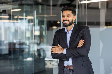 Fototapeta premium Young indian businessman with a beard smiling, looking away, and standing with crossed arms, representing confidence, success, and professional leadership in a contemporary office environment