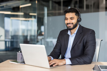Smiling man in suit jacket with headset typing on laptop at modern office desk, providing online...
