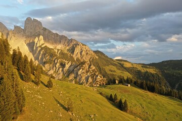 Scenic alpine valley with traditional wooden mountain huts and red rooftops, surrounded by coniferous forest. Layered mountain ridgers fade into warm evening haze at sunset. © Ivan