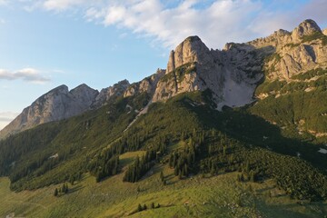 Scenic alpine valley with traditional wooden mountain huts and red rooftops, surrounded by coniferous forest. Layered mountain ridgers fade into warm evening haze at sunset. © Ivan