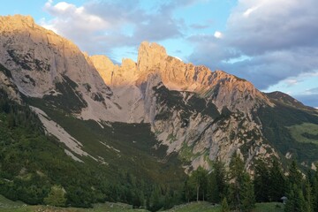 Scenic alpine valley with traditional wooden mountain huts and red rooftops, surrounded by coniferous forest. Layered mountain ridgers fade into warm evening haze at sunset. © Ivan