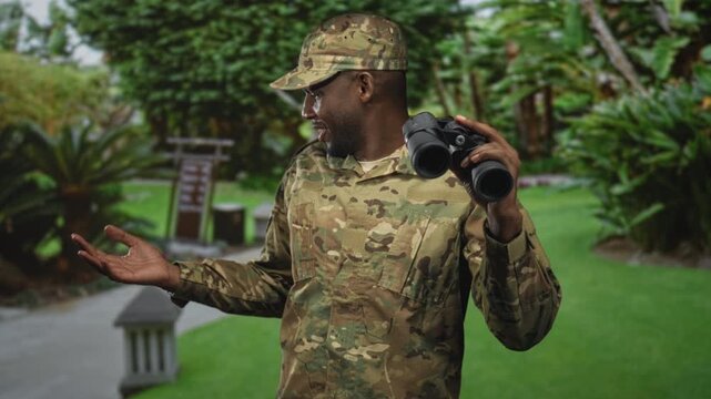 Man in military uniform smiling while holding binoculars and gesturing with open hand in forest; duty vigilance.