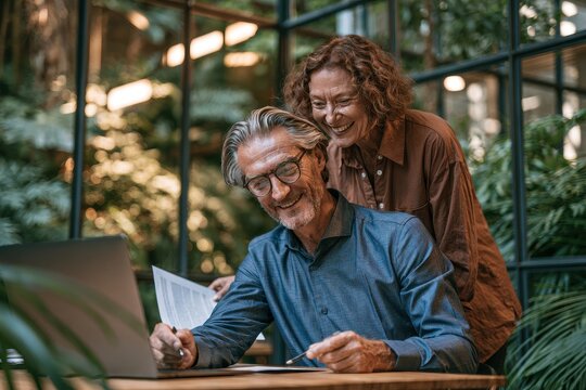 A mature couple working together in a bright, plantfilled office, with a man writing on documents and a woman smiling and looking over his shoulder