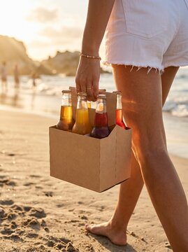 Person walking barefoot on a sandy beach at sunset, carrying a six-pack of bottled drinks.