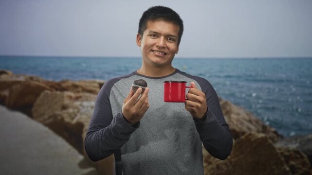 Man holding red mug and chocolate muffin in studio, smiling while posing and tasting snack; relaxation breakfast.