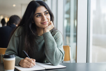 Obraz premium A young woman with long dark hair is sitting at a table, looking thoughtfully to the side while holding a pen over an open notebook in a bright indoor setting.