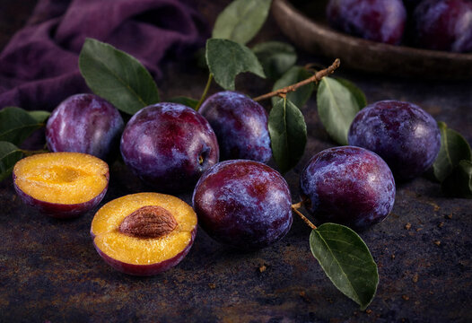 Fresh purple plums with green leaves on dark rustic background, ripe organic plum fruits, sliced plum showing yellow flesh and pit