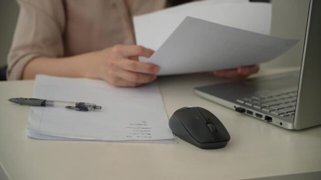 Close up footage of a woman holding and leafing through paper documents beside a laptop at an office desk. A pen, lined paper and wireless computer mouse are visible in the foreground. 