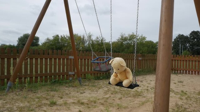 Child abduction concept with empty moving swing and teddy bear on playground. 