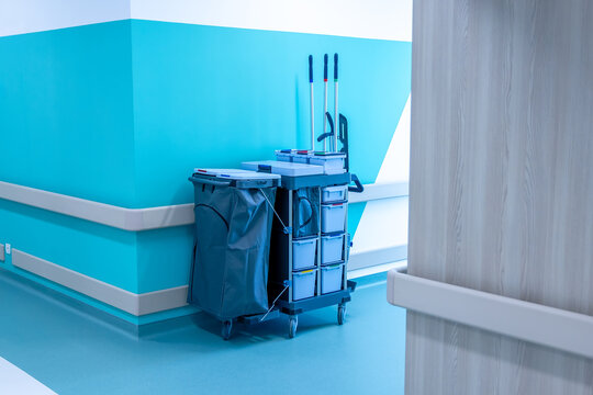 A cleaning trolley with mops, buckets and a laundry bag stands in the corner of a hospital corridor. Turquoise walls rise above the wipe clean floor. Sanitary service in a new modern hospital