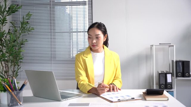Focused Professional at Work: A young woman in a bright yellow blazer diligently works at her desk, reviewing documents and typing on her laptop in a modern office setting.