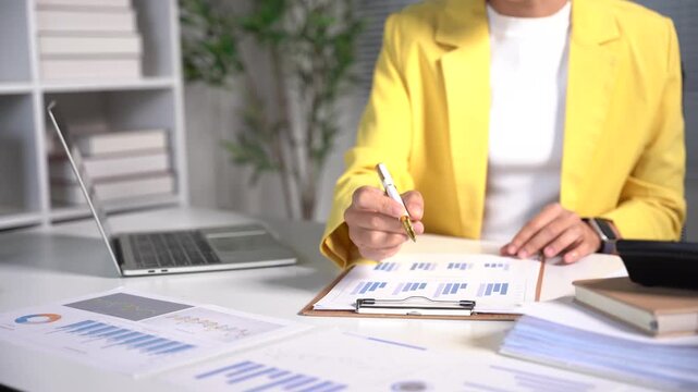 Businesswoman Analyzing Financial Report: A focused businesswoman in a vibrant yellow blazer meticulously examines charts and graphs on a clipboard, seated at her desk with a laptop and calculator.