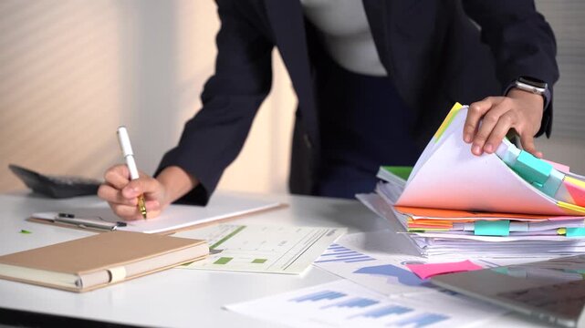 Busy Office Workspace: A person in a dark suit is actively engaged at their desk, surrounded by stacks of documents, a calculator, and a notebook, emphasizing a busy work environment.