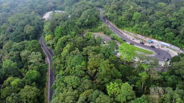 Aerial view of a winding asphalt road cutting through dense tropical forest with vehicles navigating a sharp hairpin turn in Sibolangit, North Sumatra, Indonesia.
